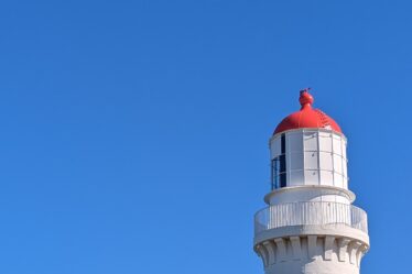 Cape Schanck Lighthouse (a tall white cylindrical building with a red top) is shown against a cloudless blue sky.