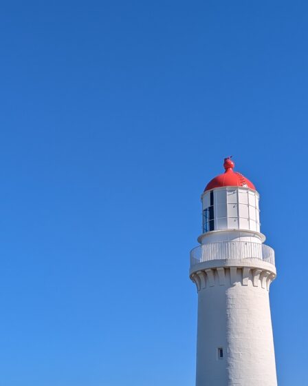 Cape Schanck Lighthouse (a tall white cylindrical building with a red top) is shown against a cloudless blue sky.